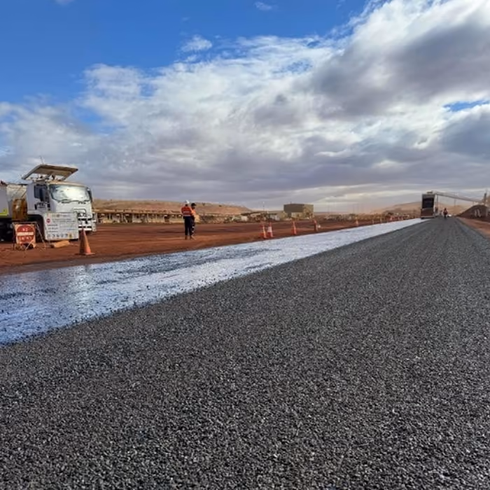 Two dump trucks and a road roller paving asphalt on a remote highway near dry hills and railway tracks.