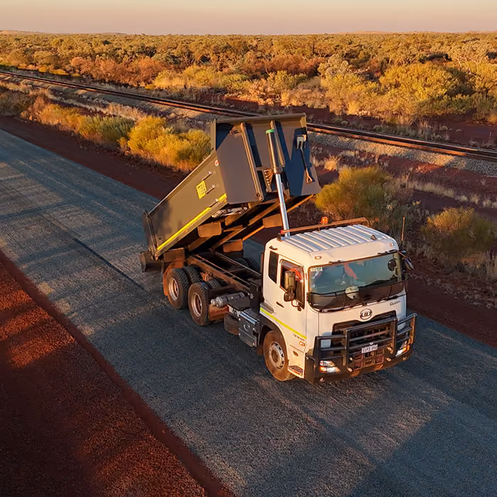 Two dump trucks and a road roller paving asphalt on a remote highway near dry hills and railway tracks.