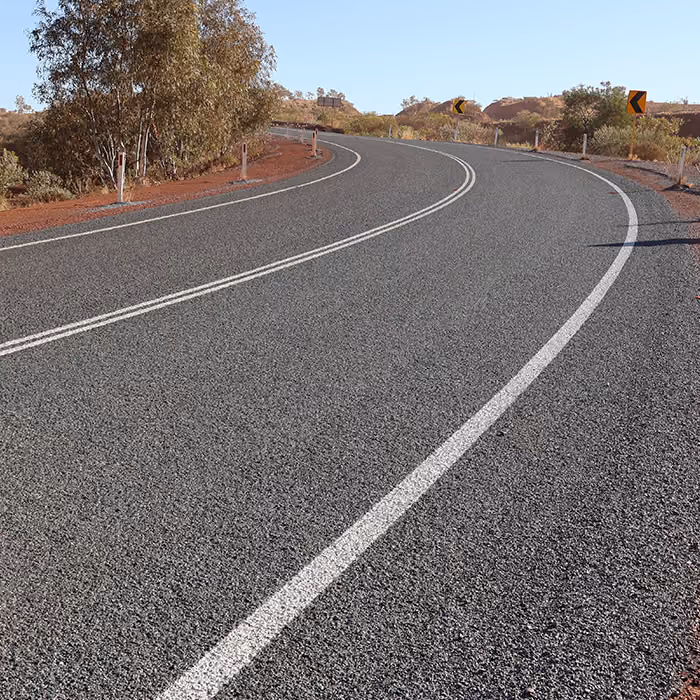 Two dump trucks and a road roller paving asphalt on a remote highway near dry hills and railway tracks.