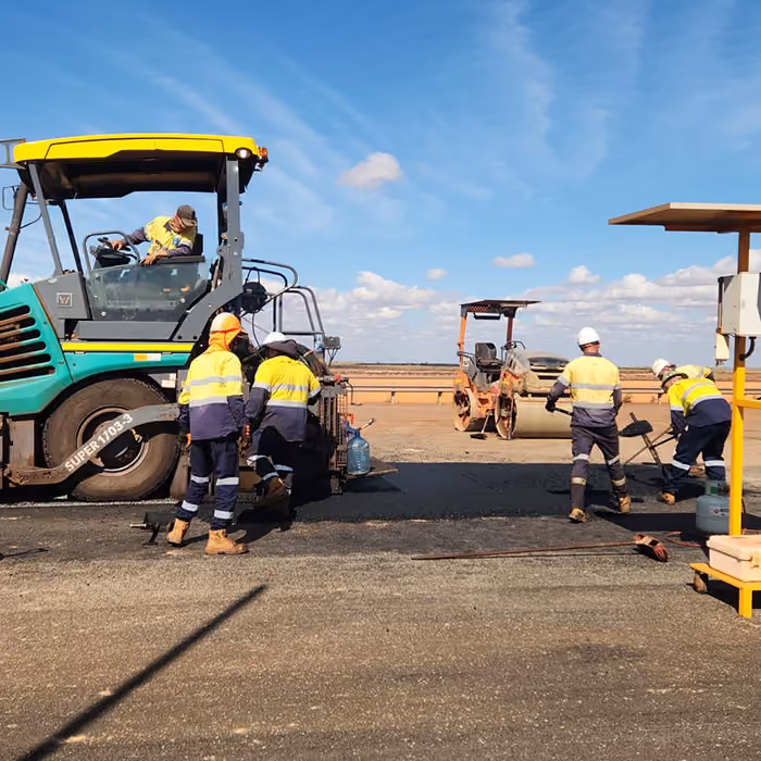 Two dump trucks and a road roller paving asphalt on a remote highway near dry hills and railway tracks.