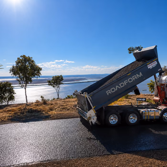 Two dump trucks and a road roller paving asphalt on a remote highway near dry hills and railway tracks.