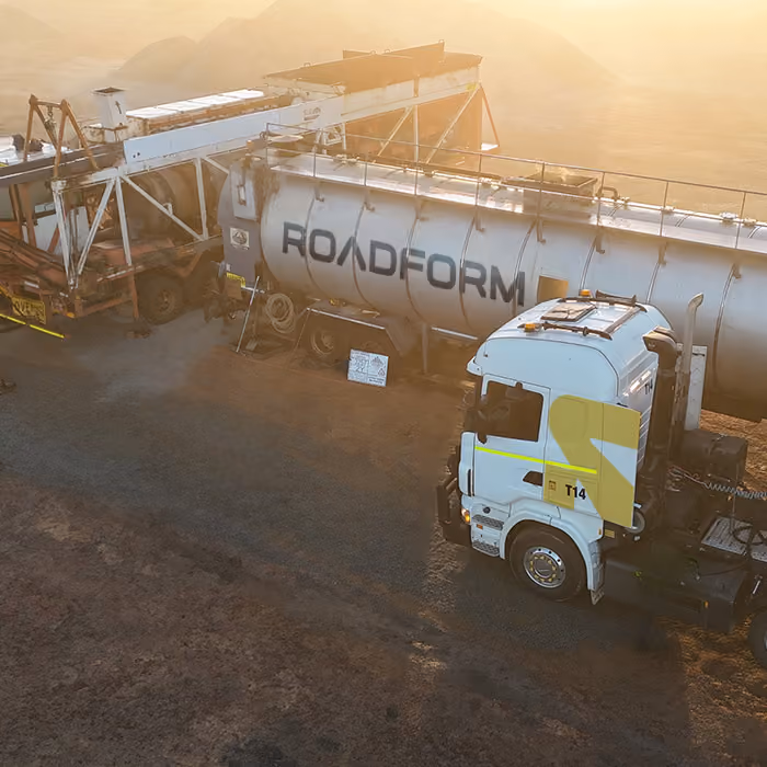 Two dump trucks and a road roller paving asphalt on a remote highway near dry hills and railway tracks.