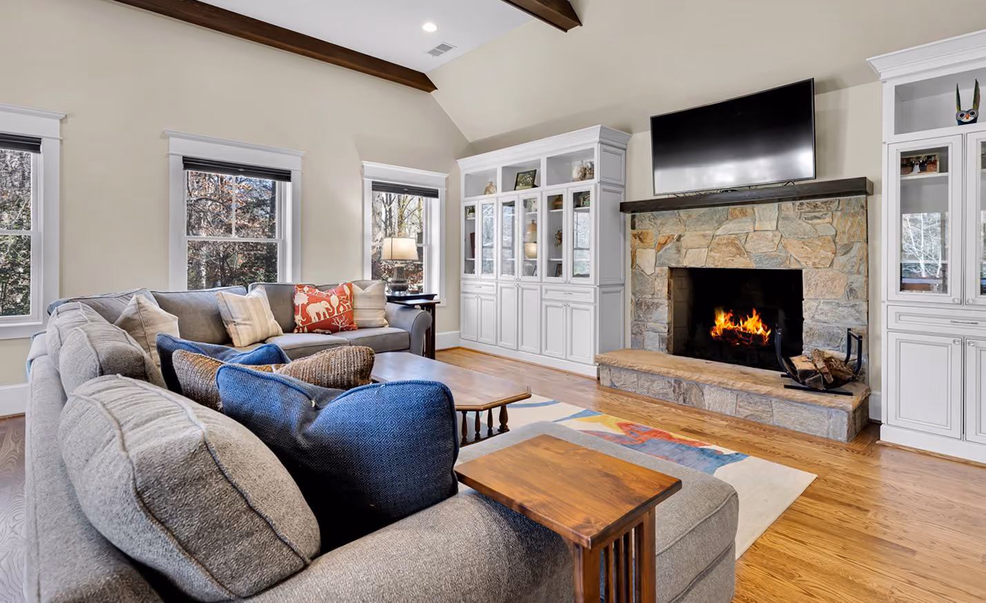 Living room with gray sectional sofa, stone fireplace with fire, mounted TV, and white cabinets.
