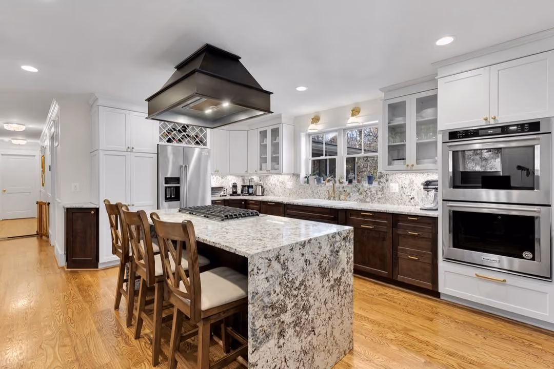 Modern kitchen with marble island, wooden chairs, stainless steel appliances, white and dark cabinets, and hardwood floor.