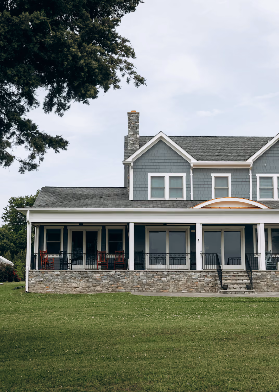 Gray two-story house with stone chimney, large porch with rocking chairs, and expansive green lawn.