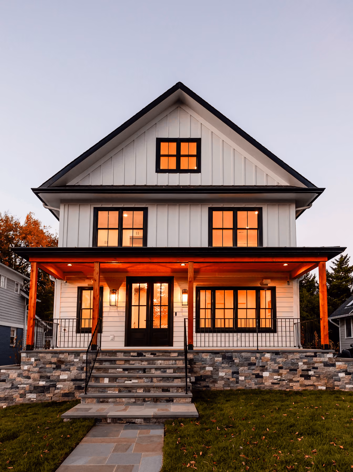 Modern two-story white house with black trim, stone foundation, front porch, and warm interior lighting.