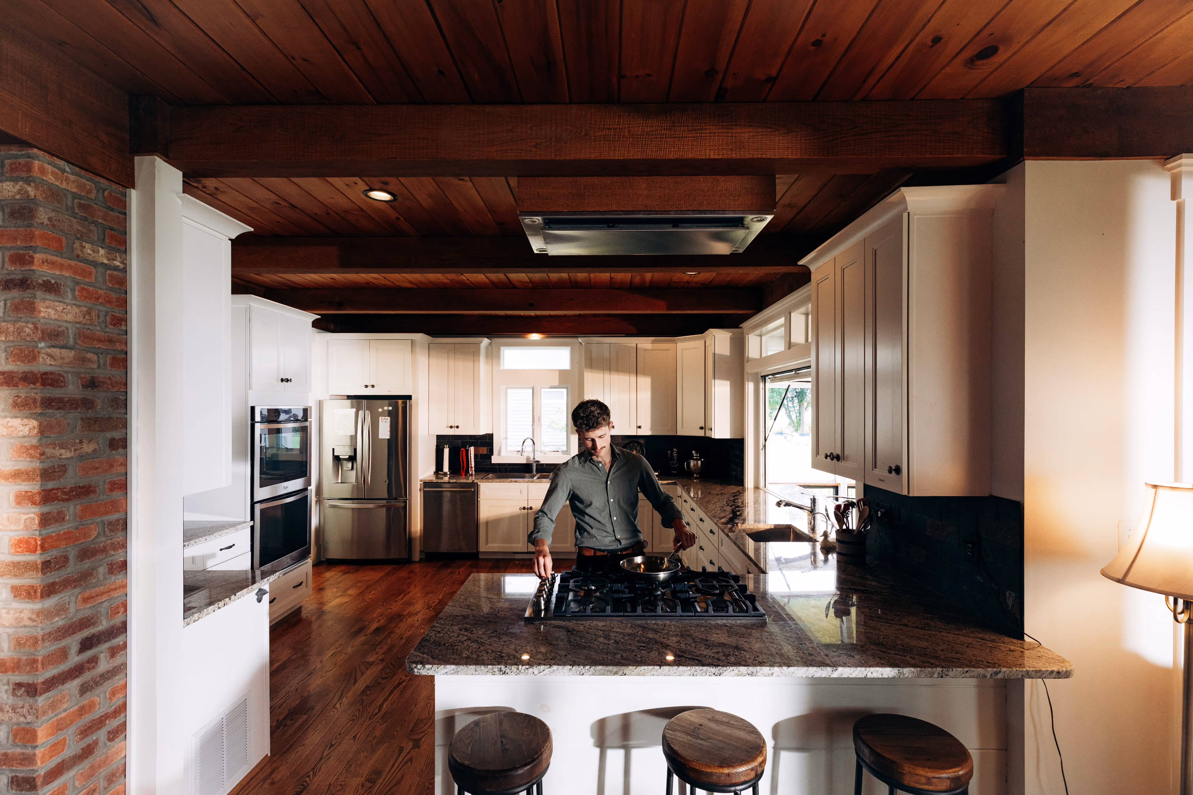 Man cooking on stove in a modern kitchen with wooden ceiling, granite counters, and white cabinets.