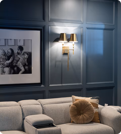 Gray sofa with round pillow under double gold wall lamps on dark paneled wall with family photo.