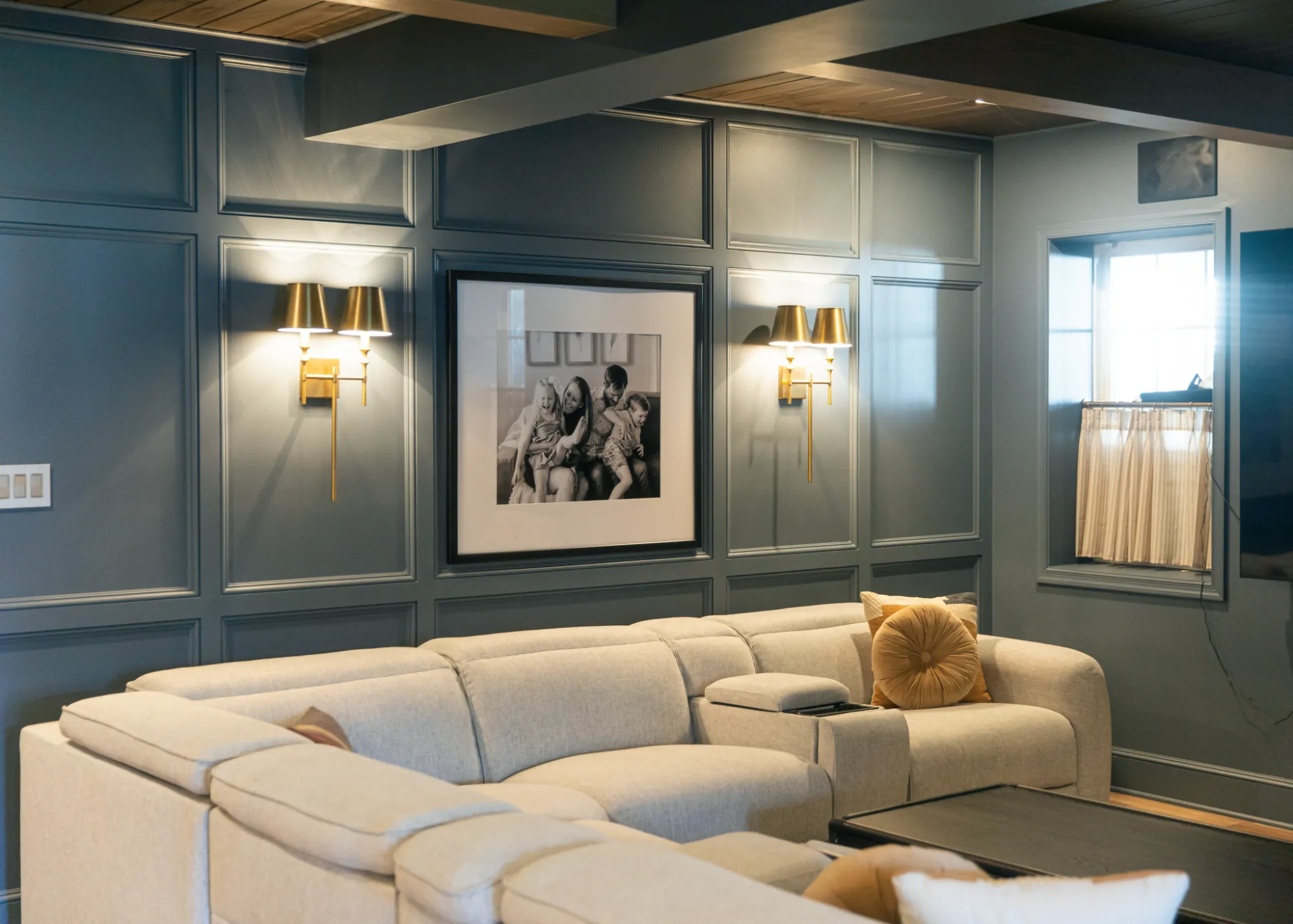 Cozy living room with beige sectional sofa, two gold wall sconces, and family photo on gray paneled wall.