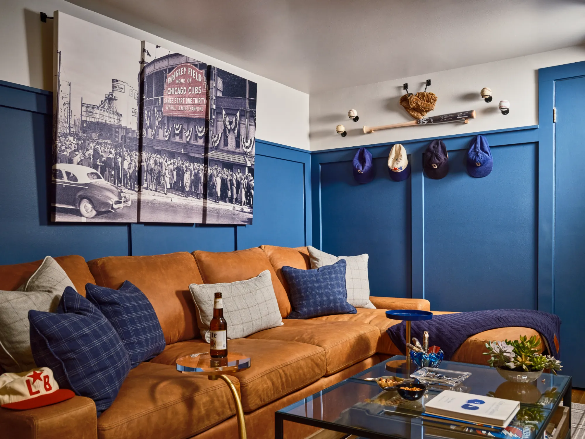 Living room with tan leather sofa, blue pillows, baseball hats, bat, glove, and vintage Chicago Cubs wall art.