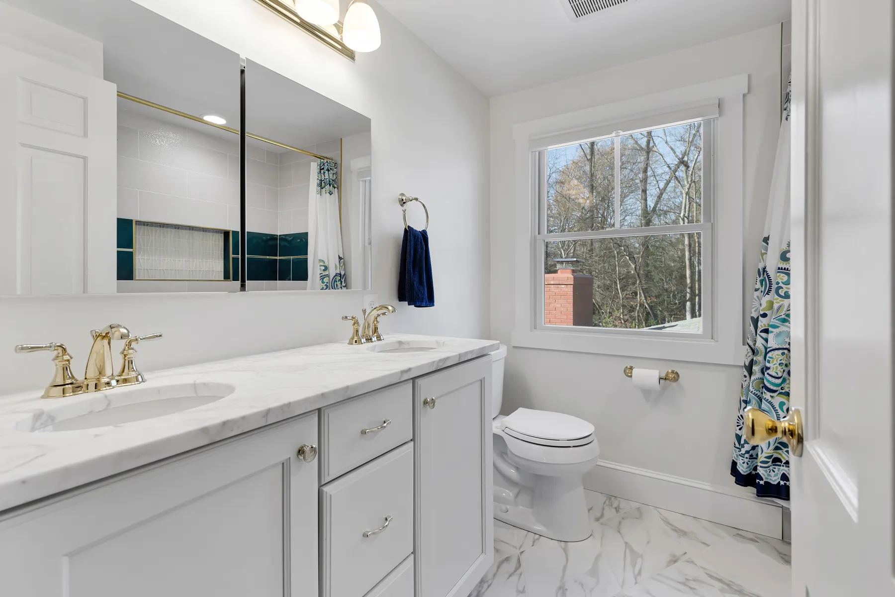 Bright bathroom with double white sinks, gold fixtures, patterned shower curtain, and window showing trees outside.