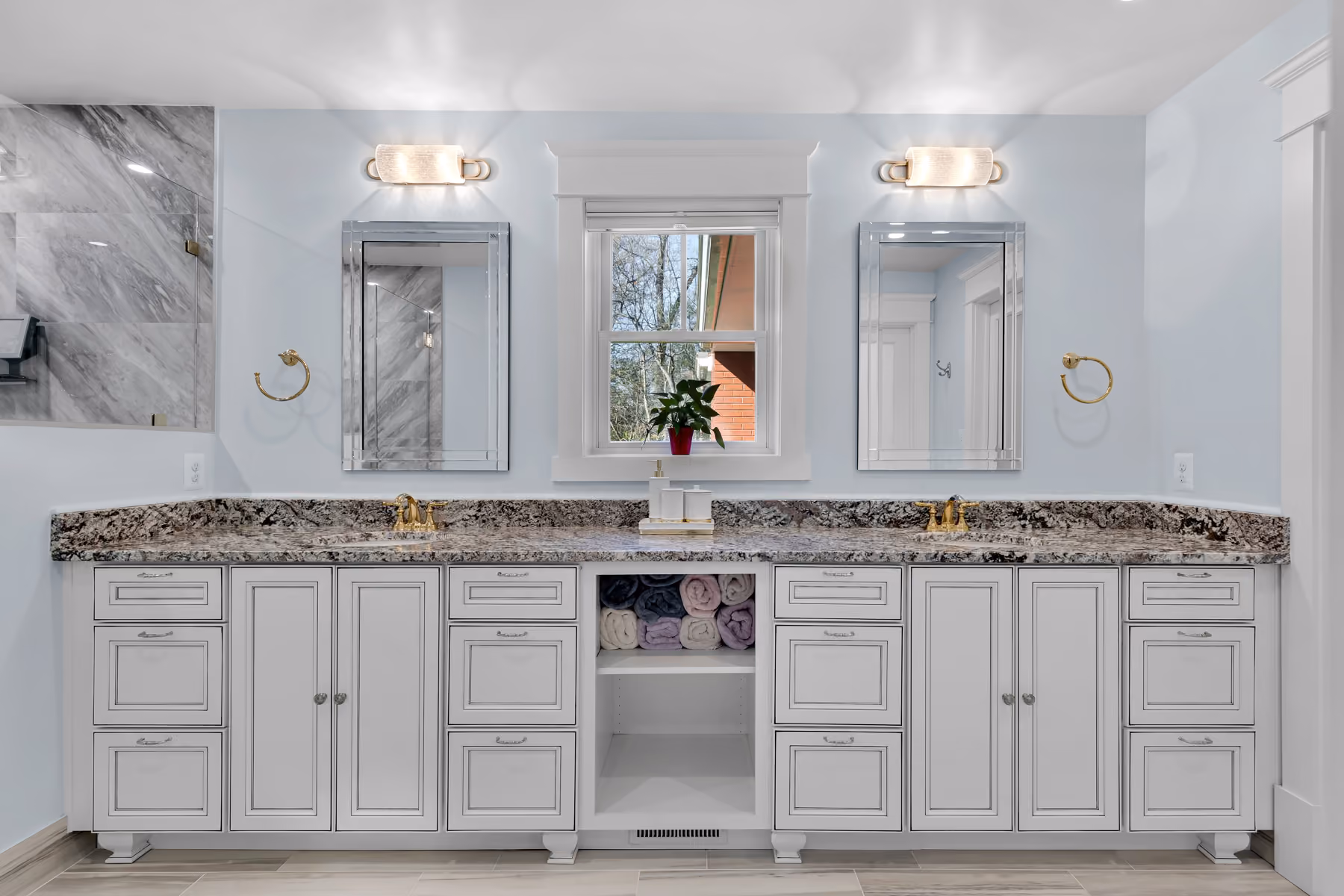 Double sink bathroom vanity with granite countertop, two mirrors, and towels in open central shelf.
