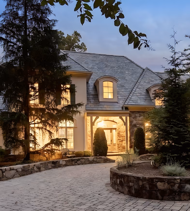 Large house with stone driveway, lit windows, and trees around during dusk.