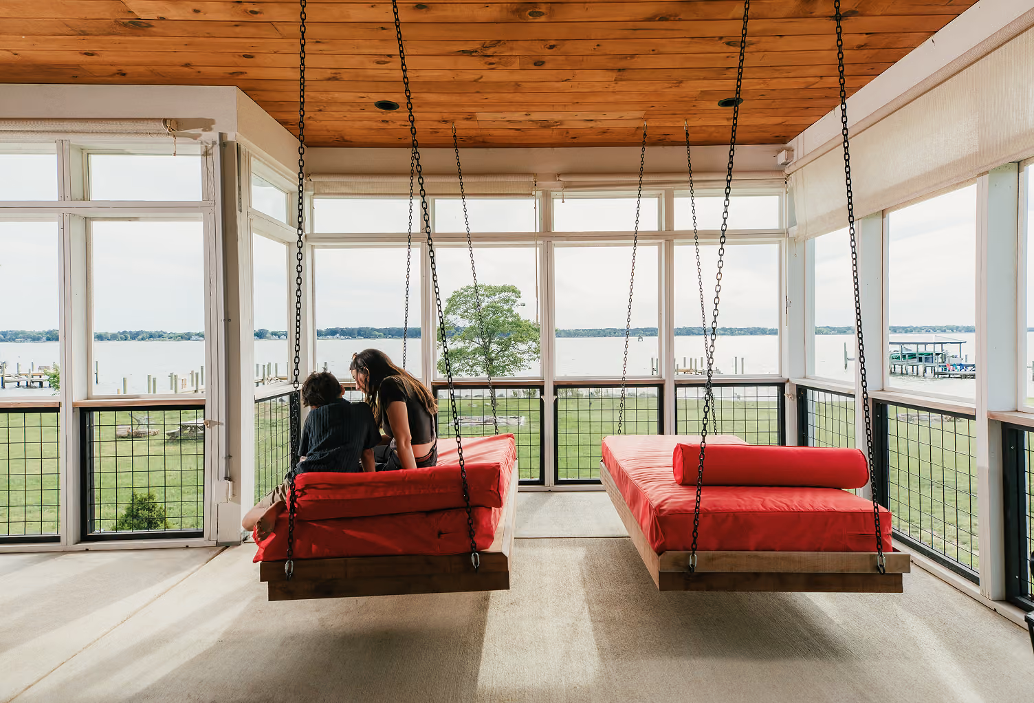 Two people sit on a red suspended swing bed in a porch overlooking a lake and grassy yard.
