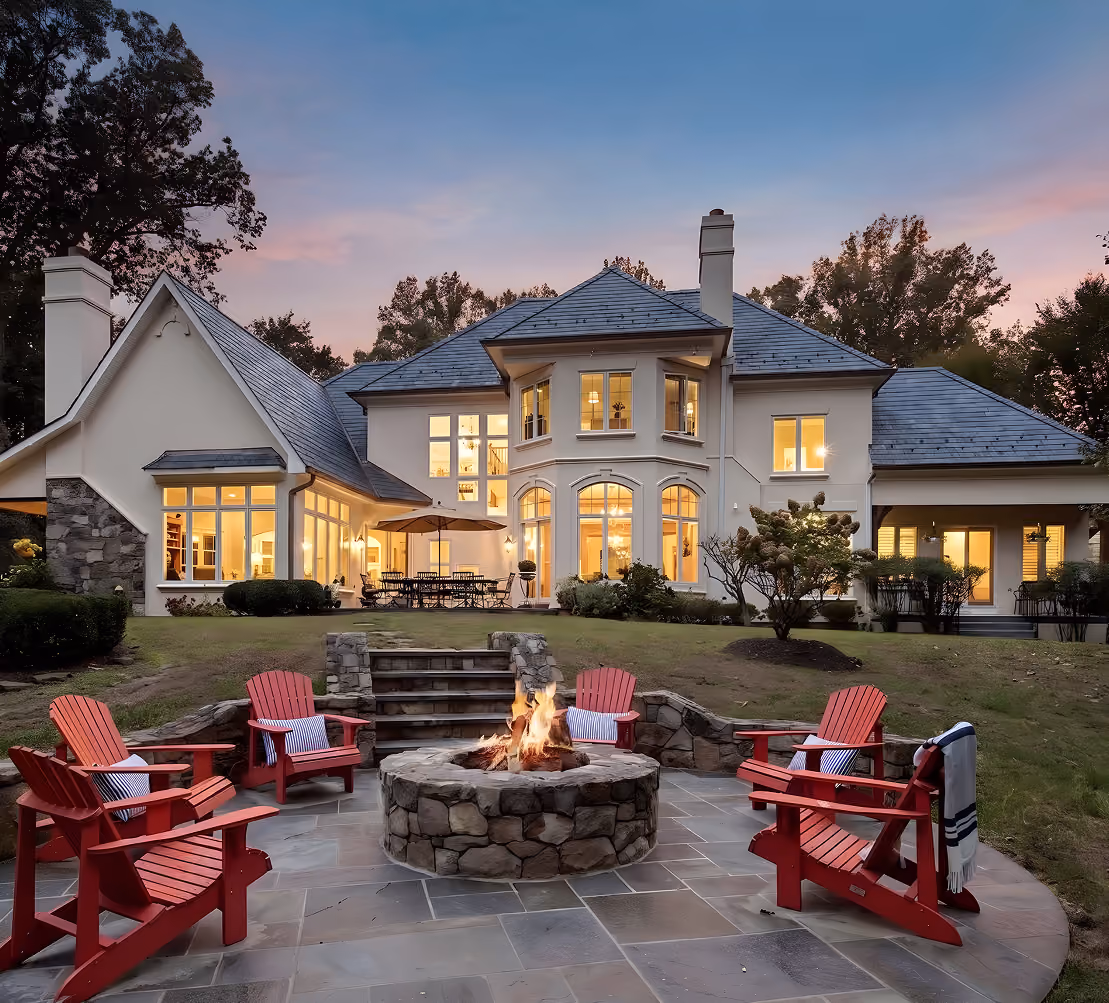 Elegant house exterior at dusk with a stone fire pit surrounded by red Adirondack chairs on patio.