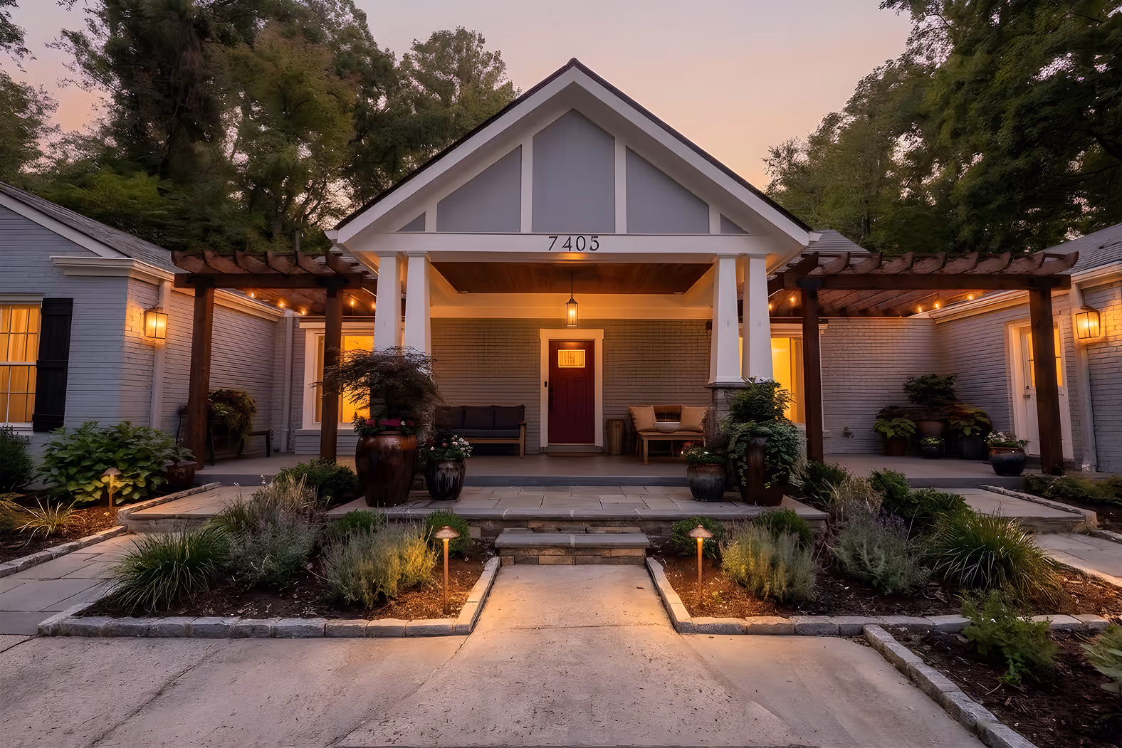 Front view of a gray house with porch, wooden pergolas, garden beds, and warm evening lighting.