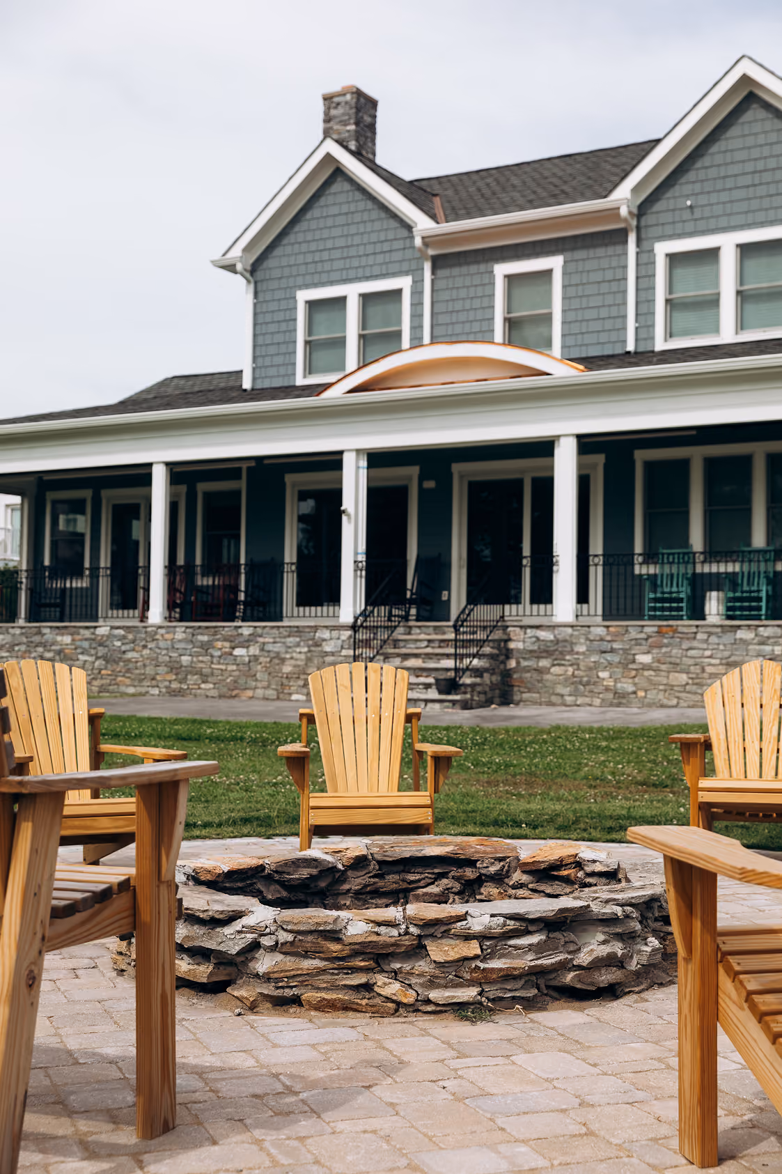 Stone firepit surrounded by wooden Adirondack chairs on a paved patio in front of a gray house.