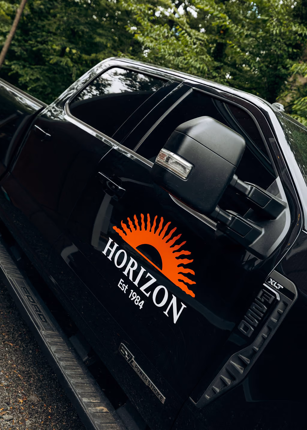 Black truck door with orange sun logo and white text reading Horizon Est 1984 parked near trees.
