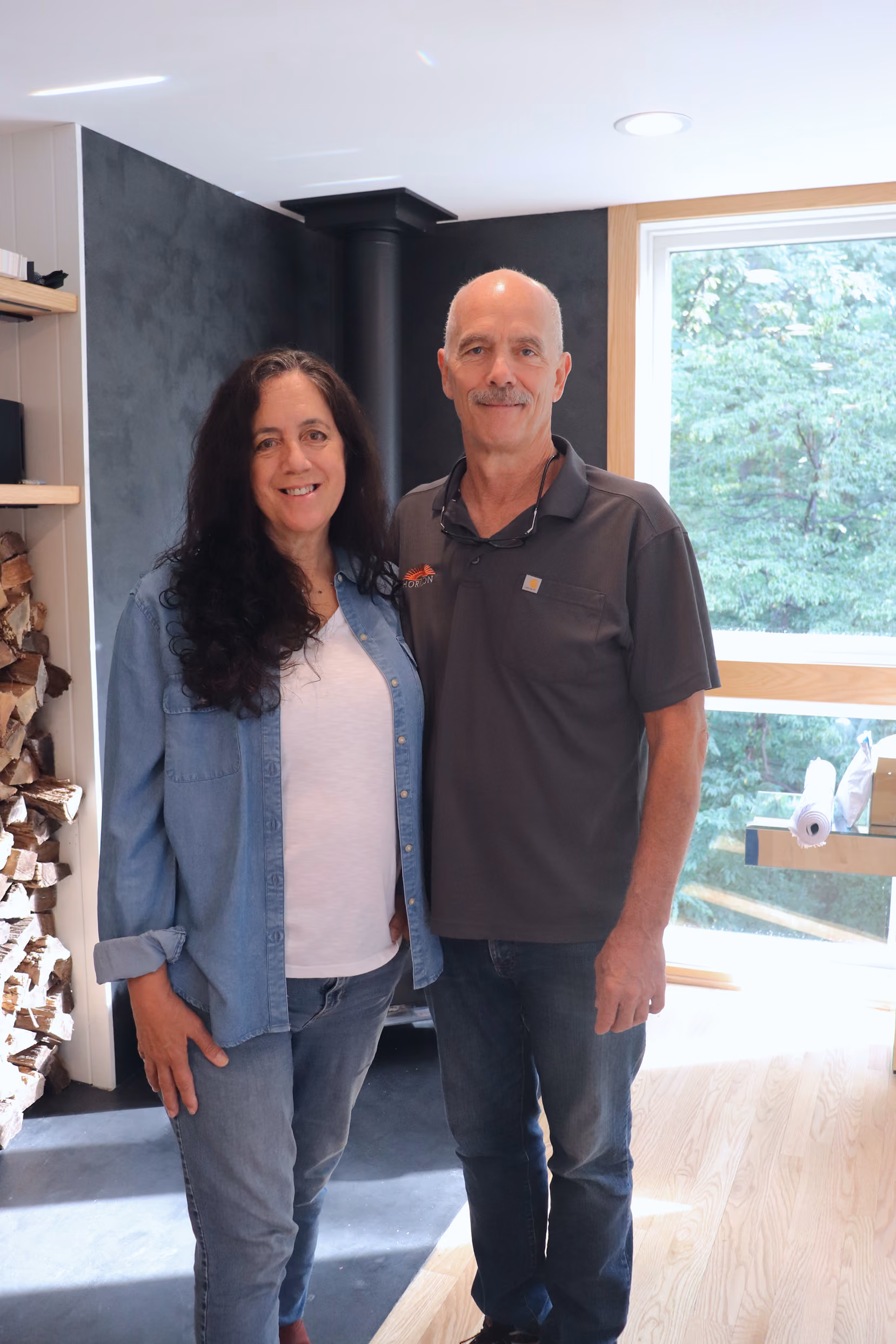 Smiling middle-aged couple standing inside a room with a wood stove and large window showing trees outside.