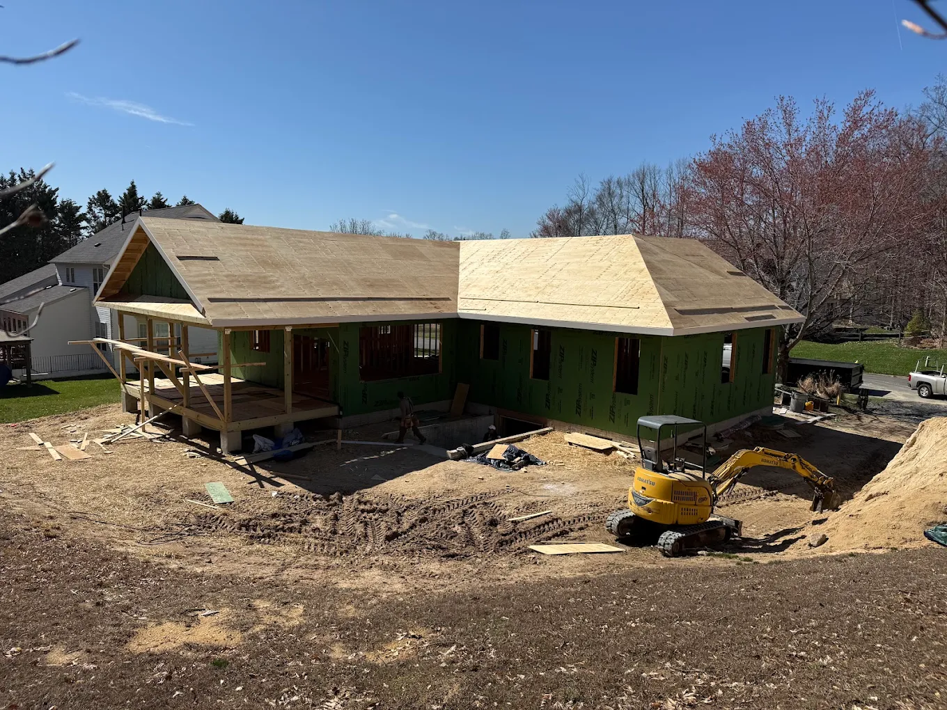 Partially framed house under construction with a yellow excavator and dirt yard around it.