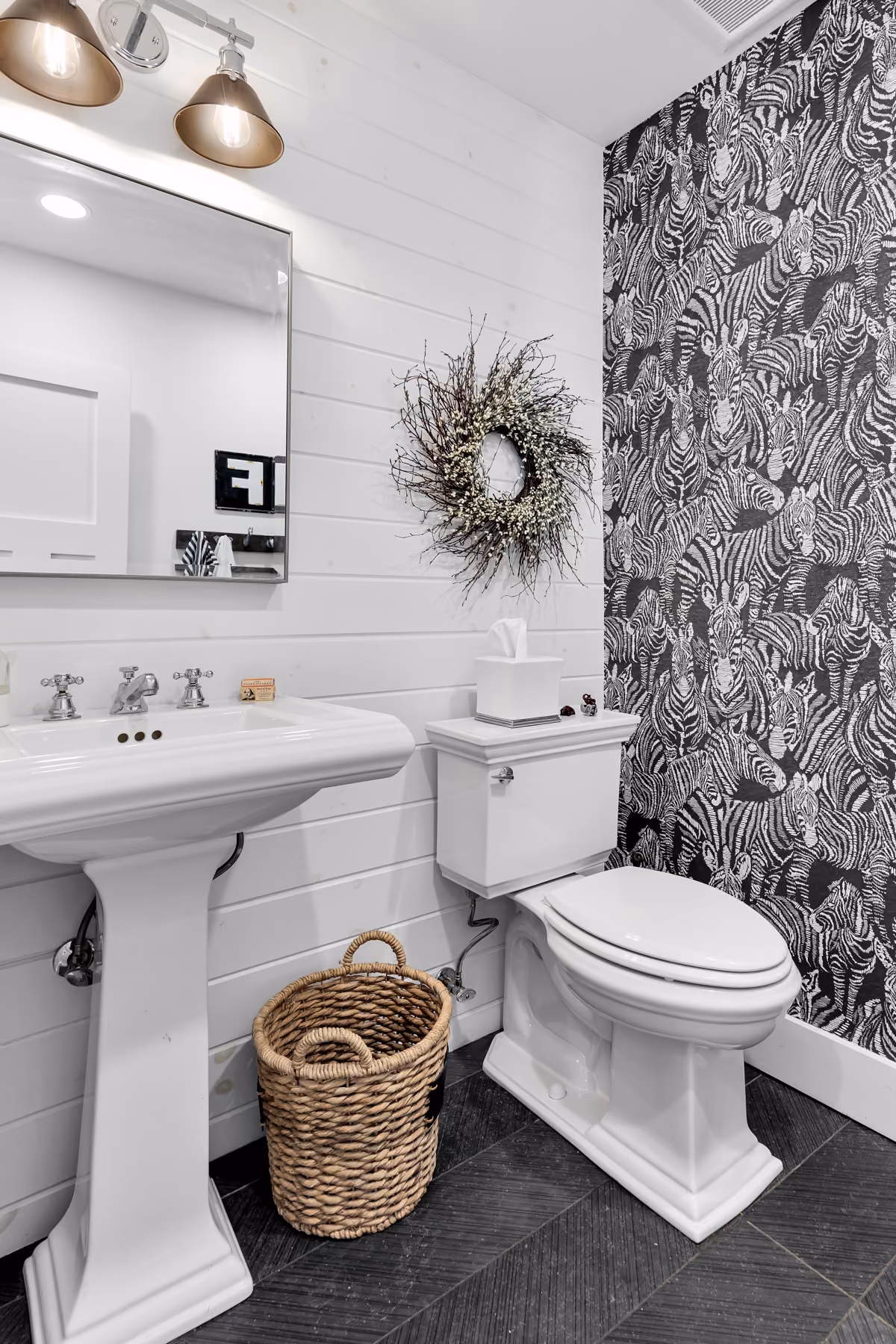 White bathroom with a pedestal sink, toilet, zebra patterned wallpaper, and a woven basket on dark floor.