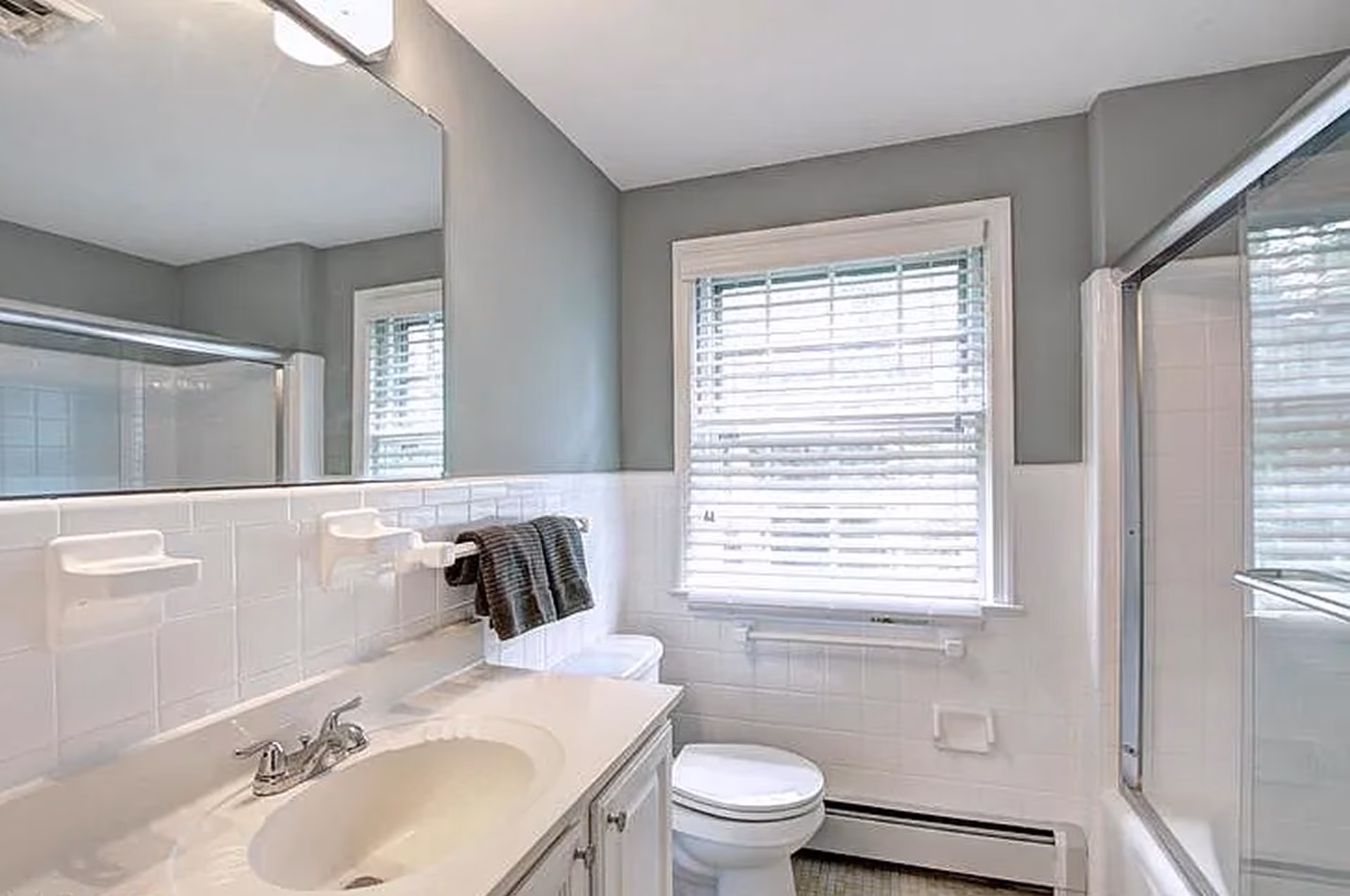Bright bathroom with white sink, toilet, large mirror, window blinds, and glass shower door.