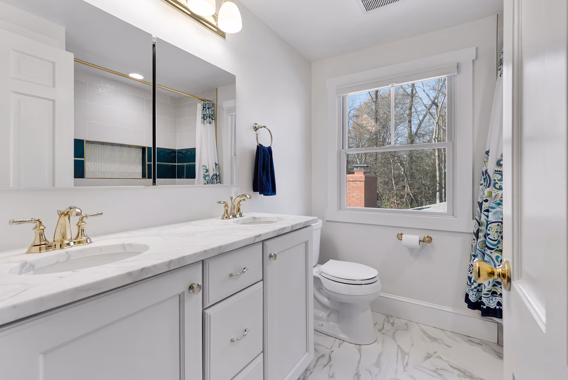 Bright bathroom with white double sink vanity, gold faucets, marble floor, window, and toilet.