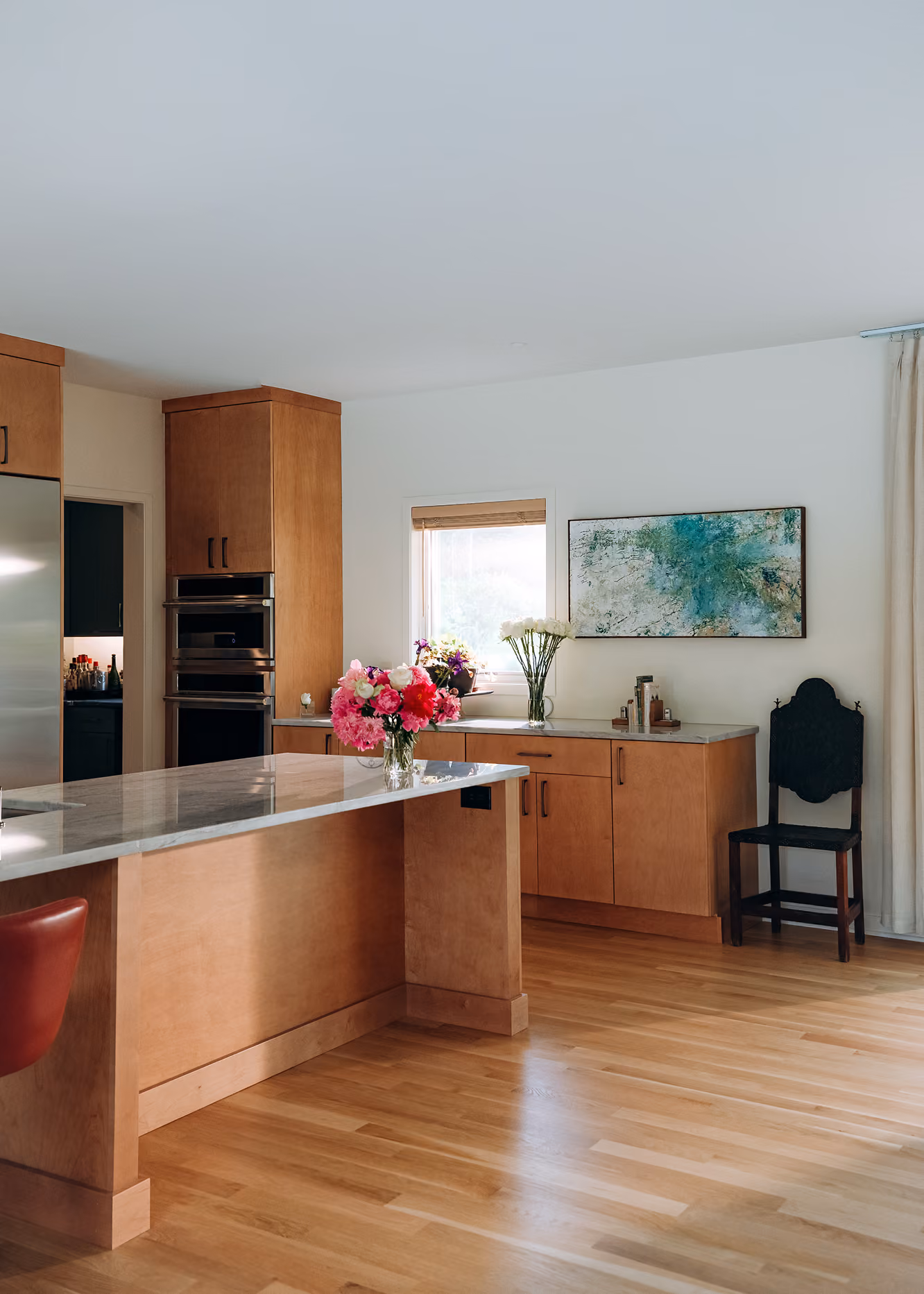 Modern kitchen with wooden cabinets, marble island, flowers on counters, a window, and a black wooden chair.