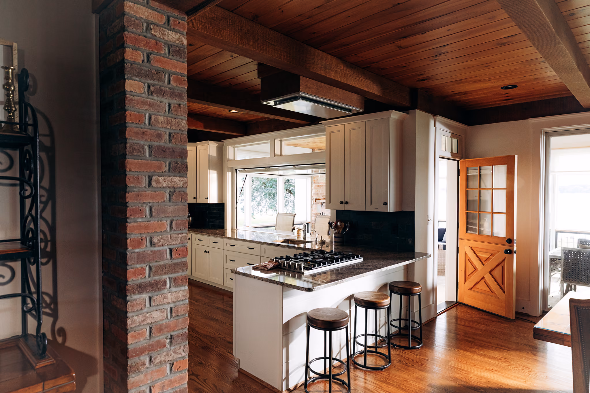 Kitchen with wood ceiling, brick column, white cabinets, granite island, and three stools near open wooden door.