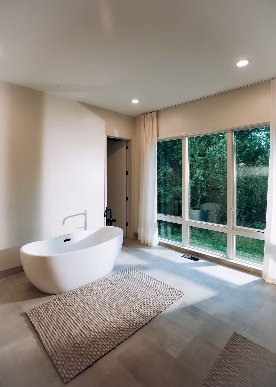 Bright modern bathroom with large window, white freestanding tub, and beige rug on tile floor.