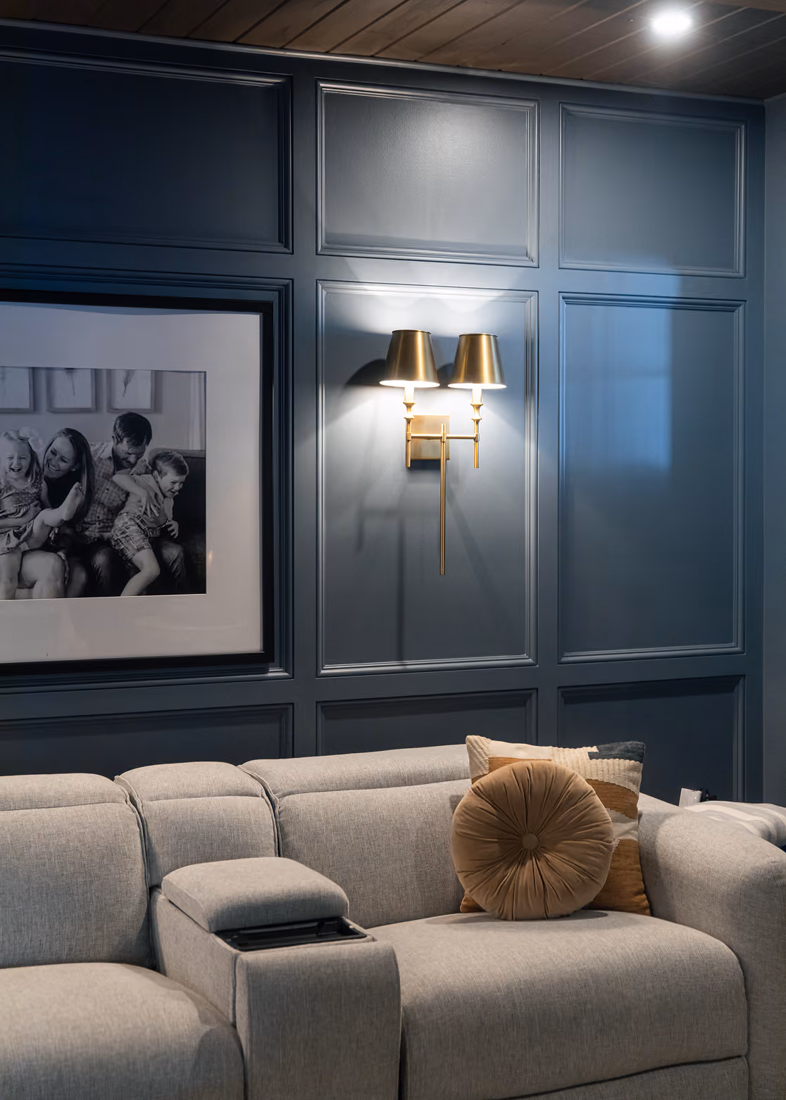 Gray sectional sofa with decorative pillows under two lit brass wall sconces on paneled blue wall.