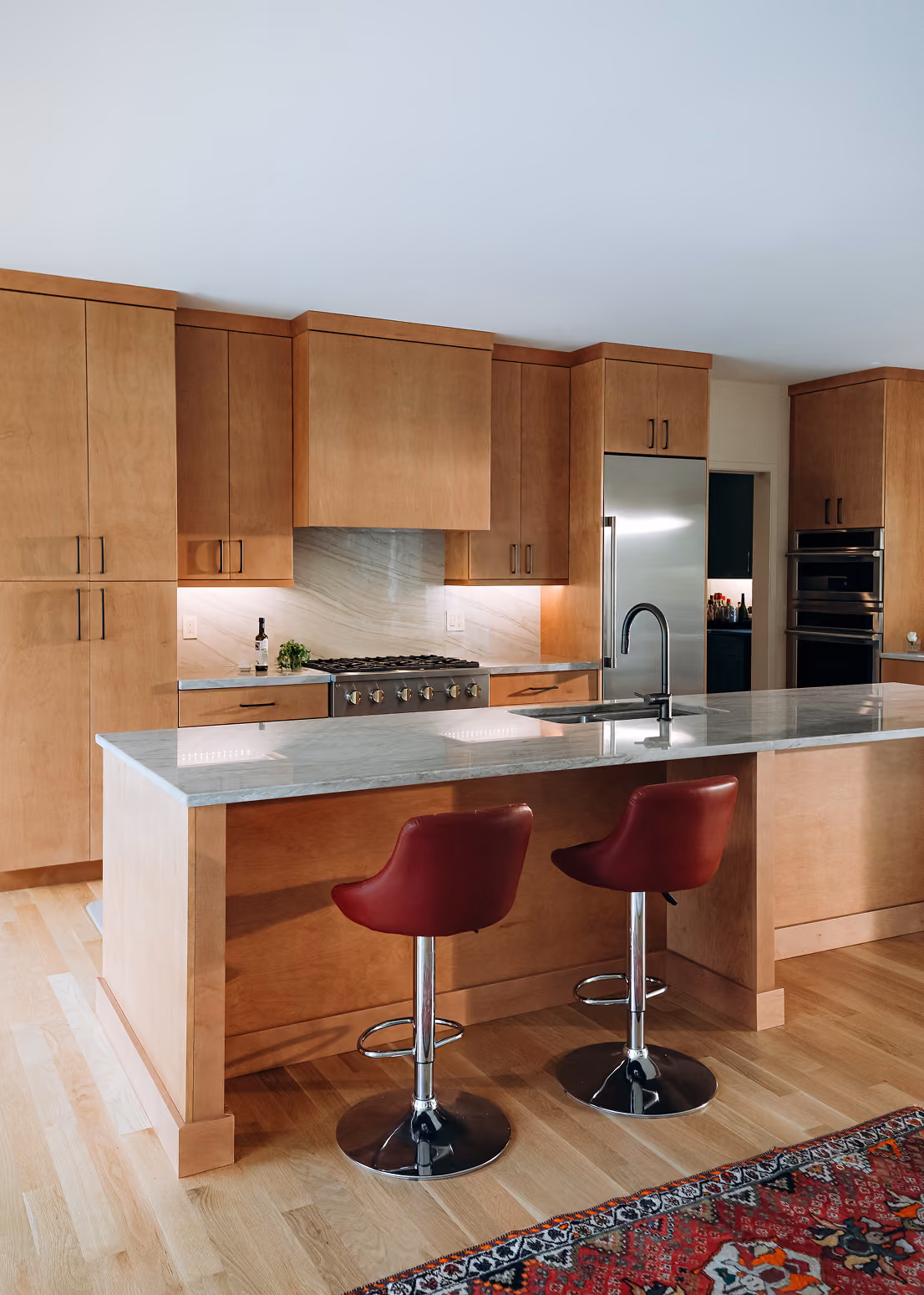 Modern kitchen with wooden cabinets, marble island, two red bar stools, and stainless steel appliances.