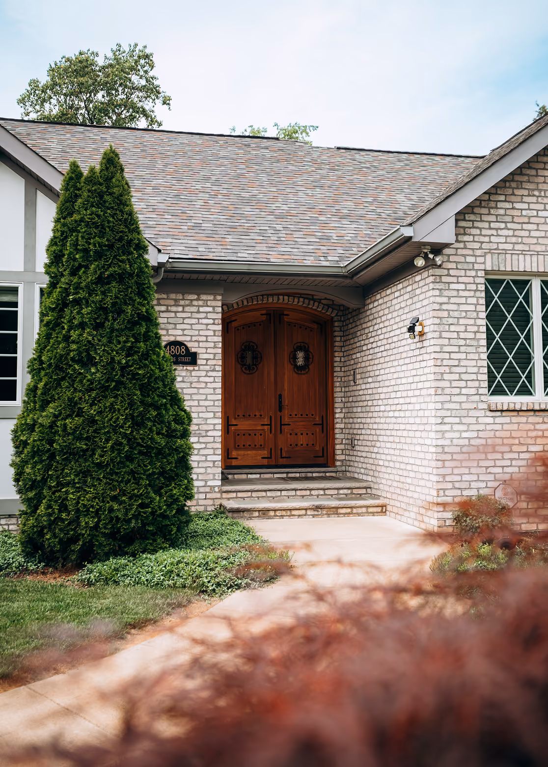 Front entrance of a brick house with a wooden double door and a tall evergreen shrub.