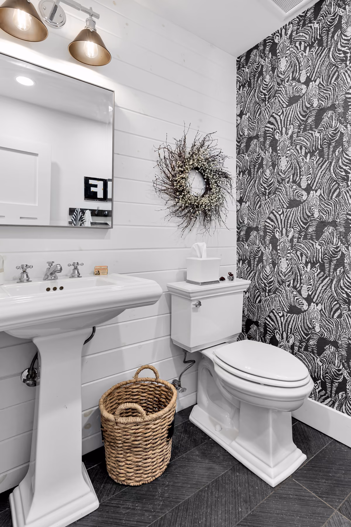White bathroom with zebra-patterned wallpaper, pedestal sink, toilet, woven basket, and decorative wreath.