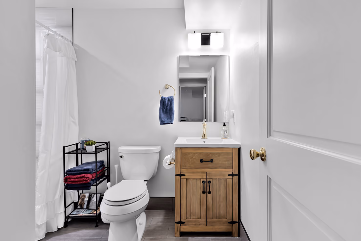 Bright bathroom with white toilet, wooden vanity, mirror, towel rack, and black shelf with towels and decor.