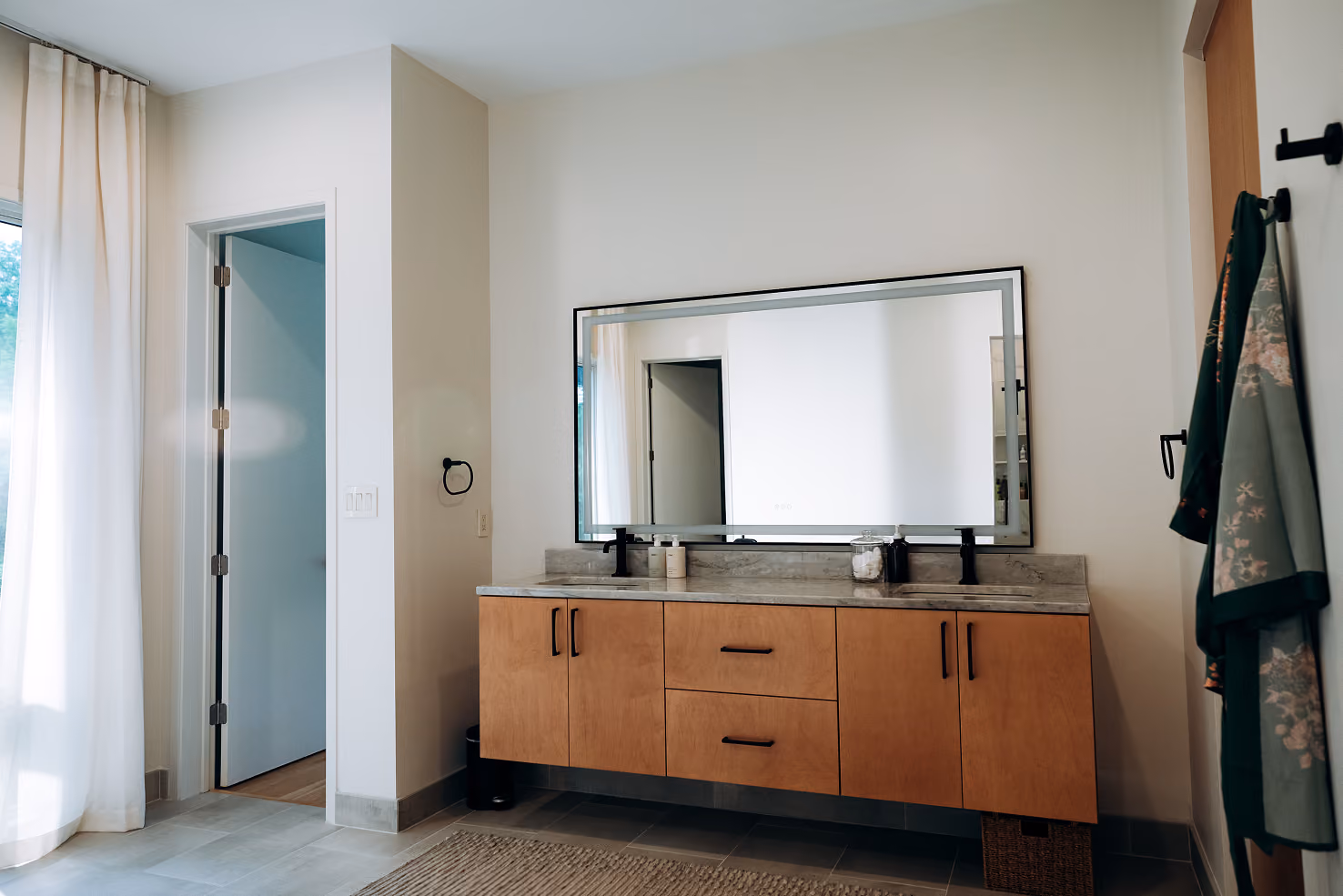 Modern bathroom with double sink vanity, large mirror, and robes hanging on wall hooks.