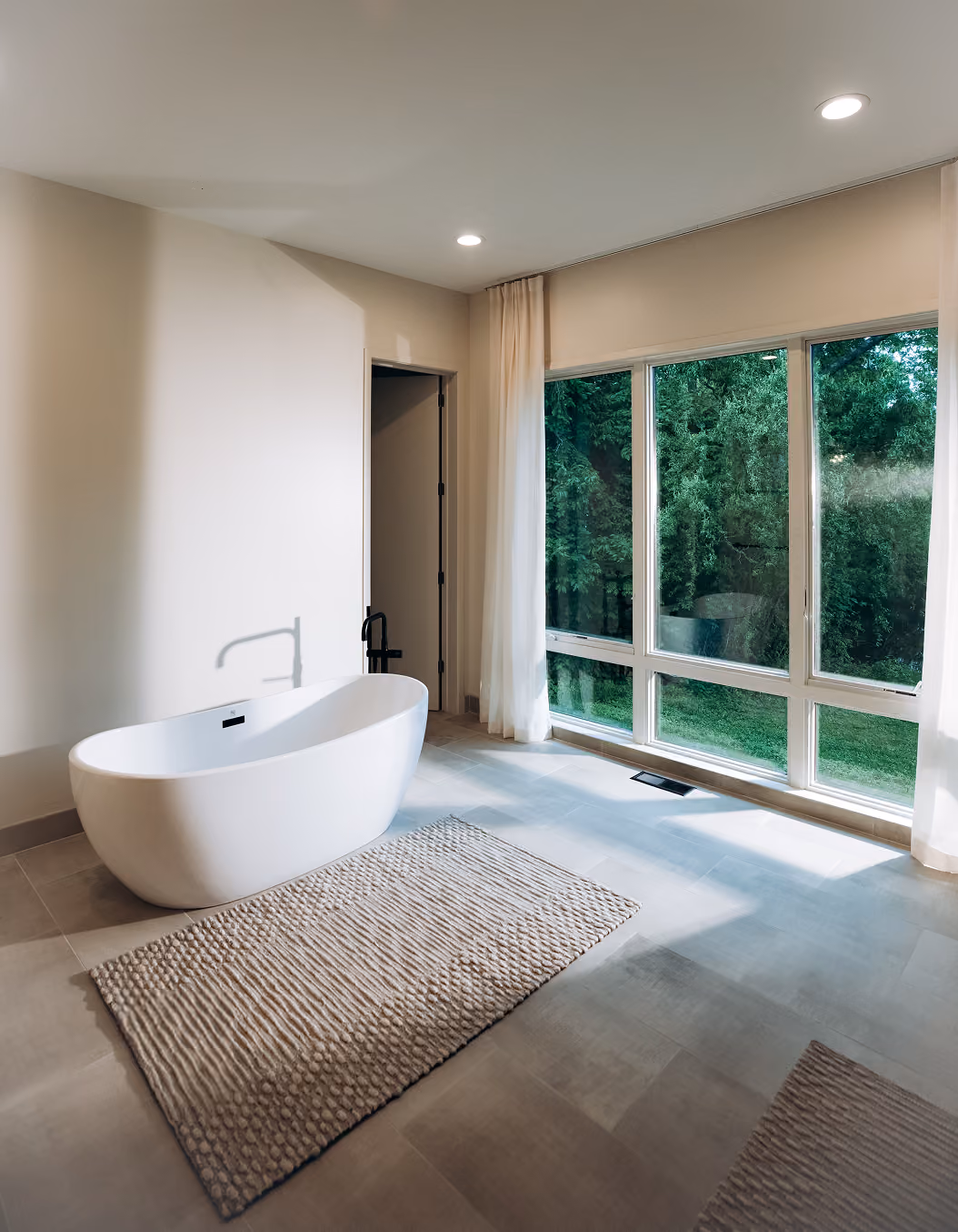 Modern bathroom with a white freestanding tub, beige rug, and large windows showing green trees outside.
