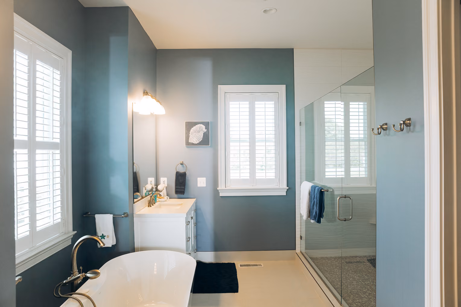 Modern bathroom with white bathtub, vanity, glass shower, blue walls, and white shuttered windows.