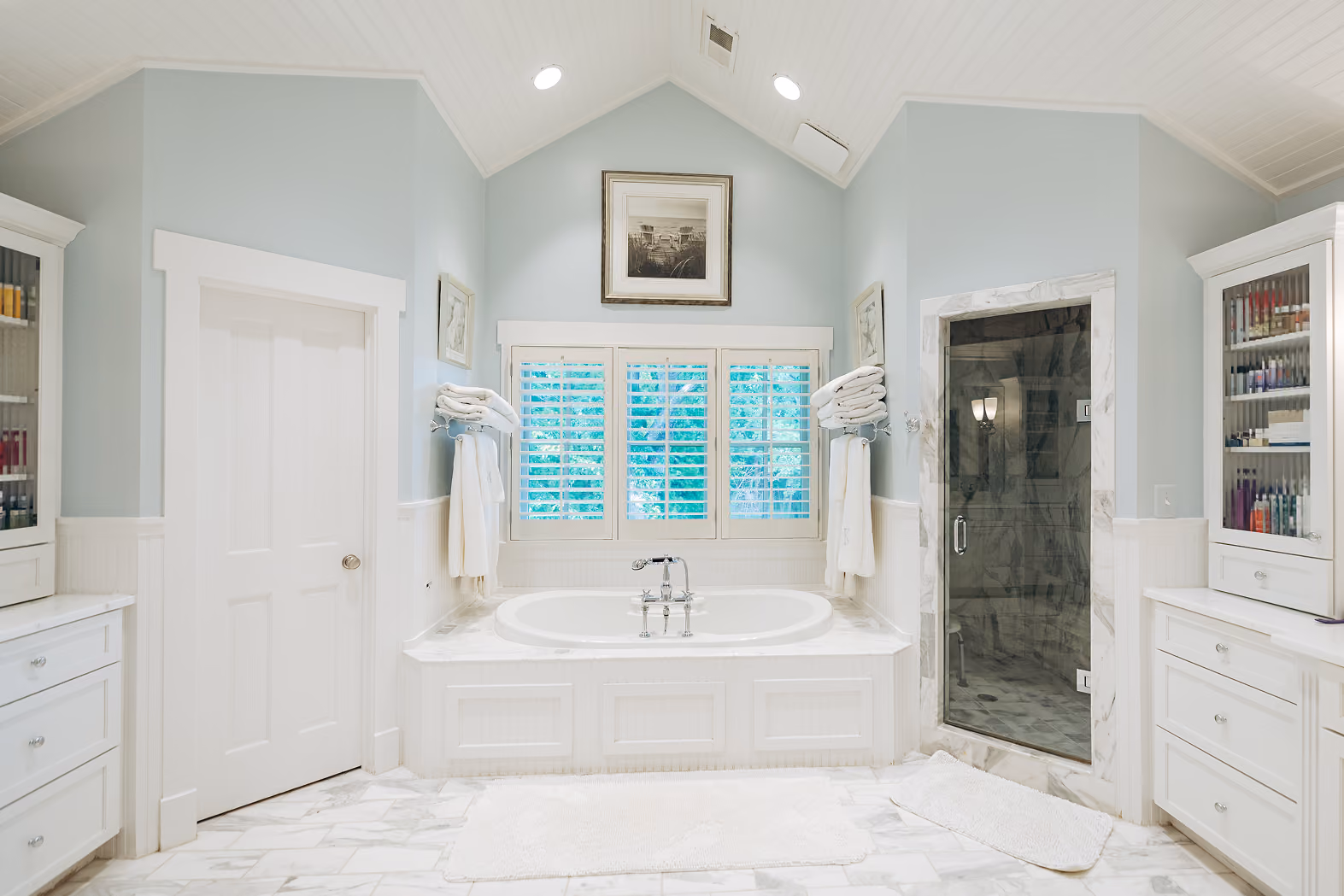 Bright bathroom with white bathtub, marble floor, glass shower, white cabinets, and windows with shutters.