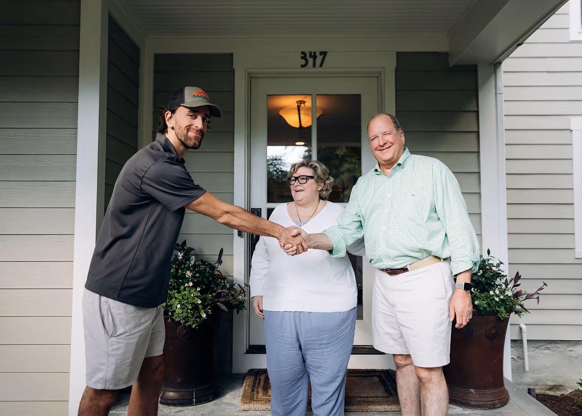 Man in a cap shaking hands with another man outside a house, with a woman smiling between them.