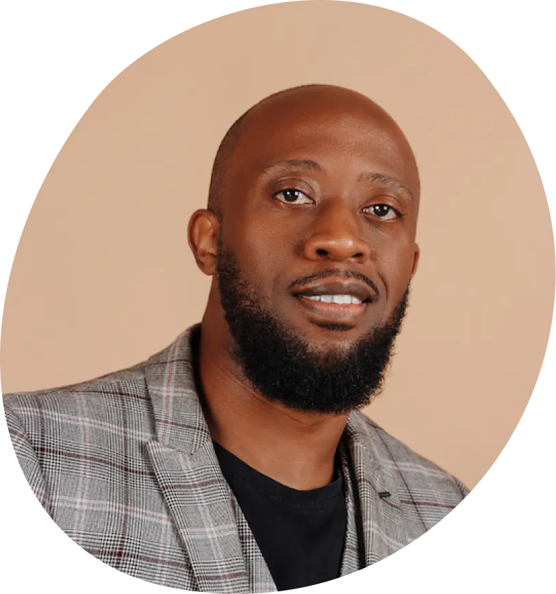 Bearded man wearing a gray plaid blazer and black shirt, smiling against a beige background.