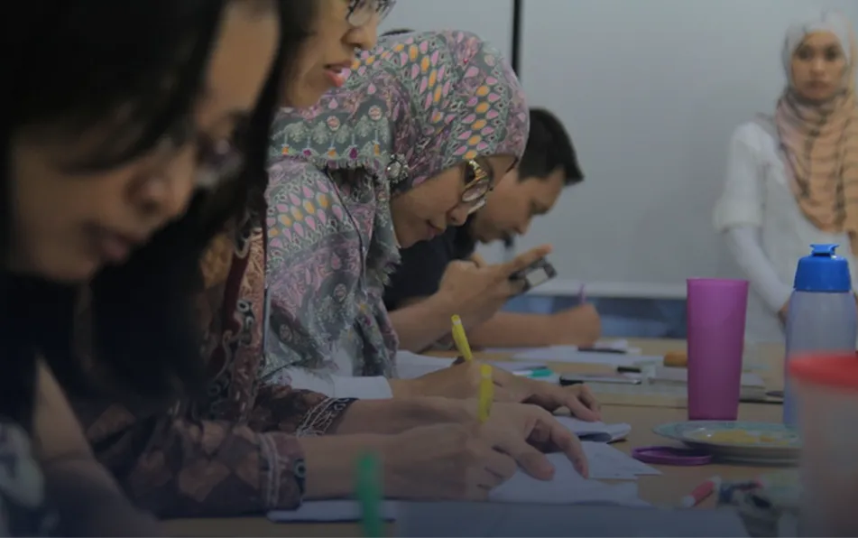 People seated at a table writing in notebooks, one person using a smartphone, focus on woman with patterned hijab.