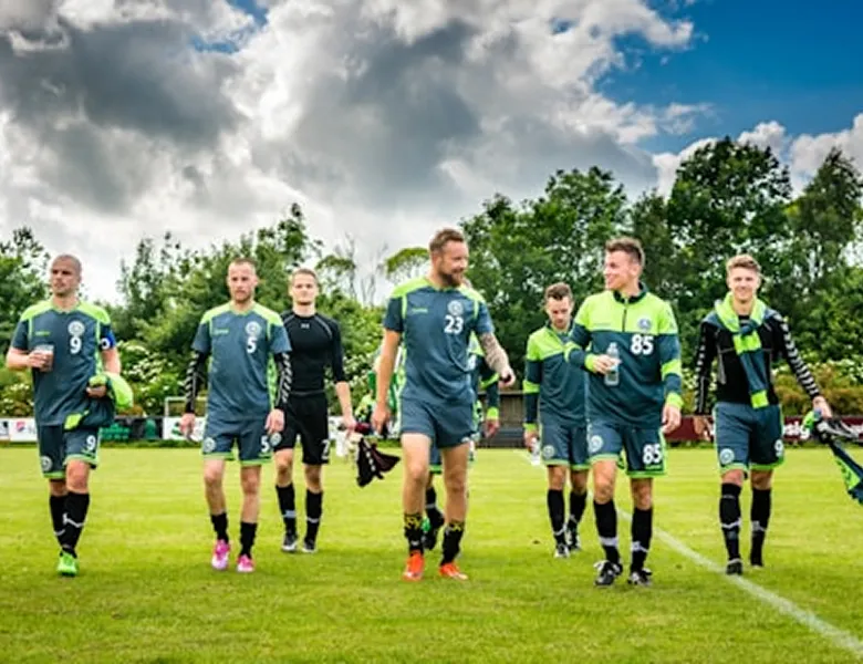 Six male soccer players in dark green uniforms walking on grass under a cloudy sky.