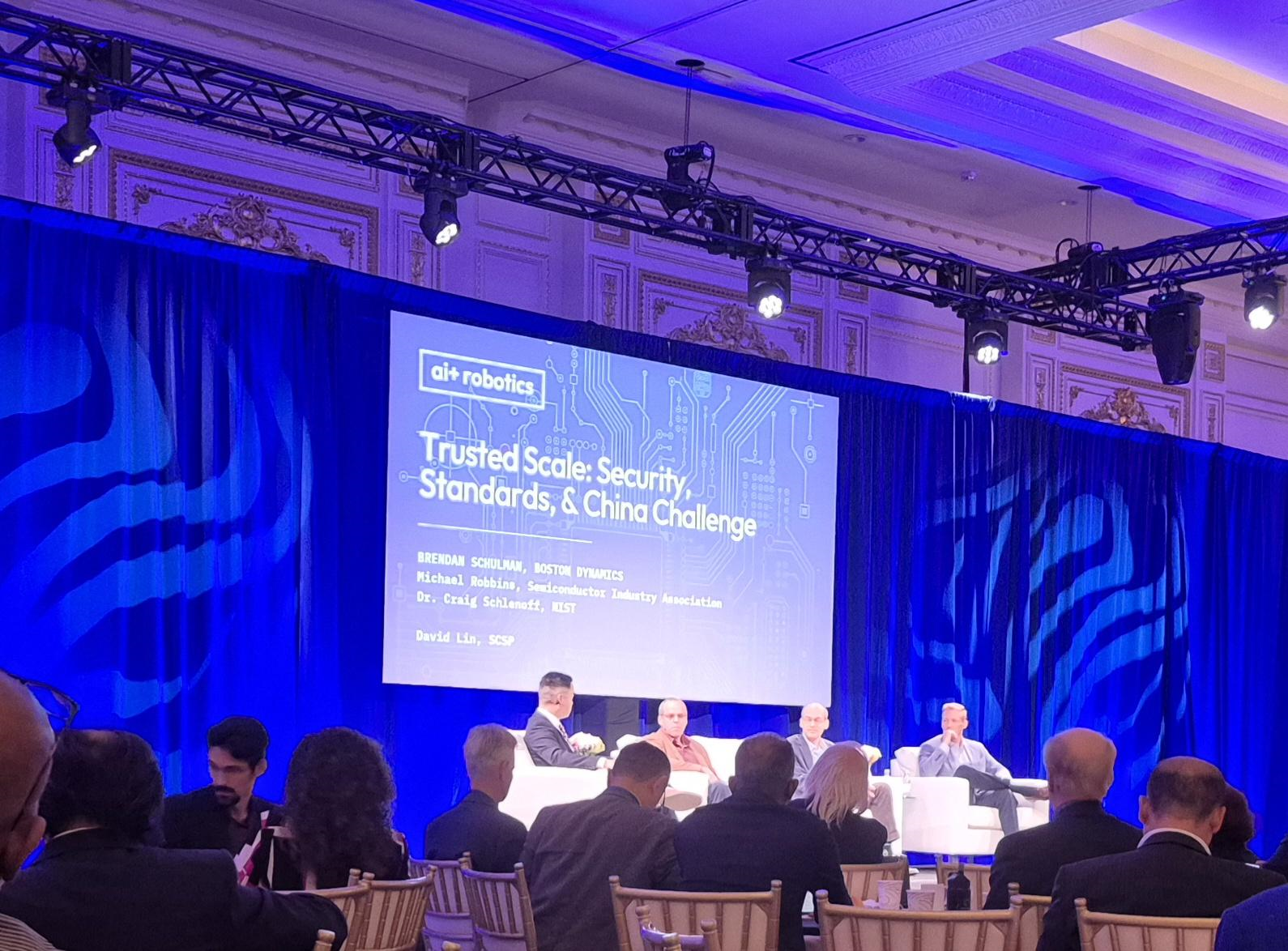 Panelists seated onstage beneath a screen reading “Trusted Scale: Security, Standards, & China Challenge” at the AI + Robotics conference.