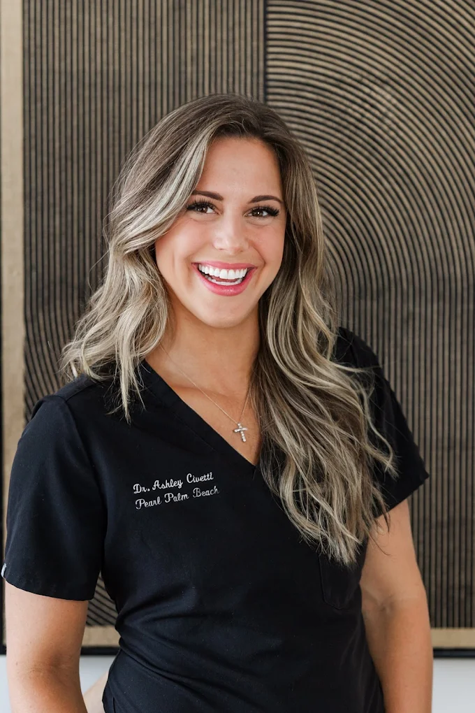 Dr. Ashley Civetti smiles warmly in black Pearl Palm Beach Dental scrubs, wearing a silver cross necklace in front of a textured neutral wall.