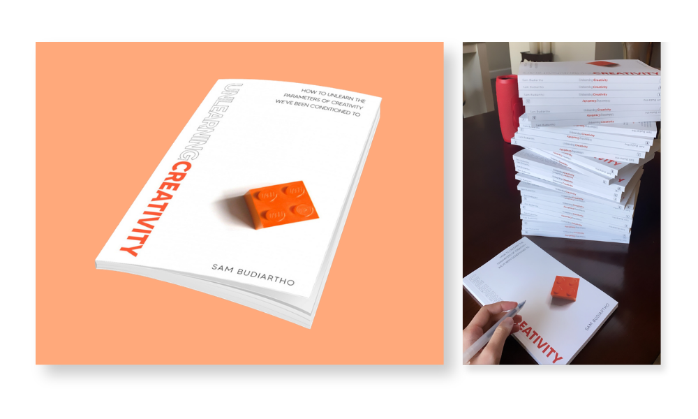 Two images showing the book 'Unlearning Creativity' by Sam Budiartho; the left image shows the book cover with an orange Lego brick on it over a peach background, the right image shows a hand holding a pen above the same book on a table alongside a tall stack of identical books.