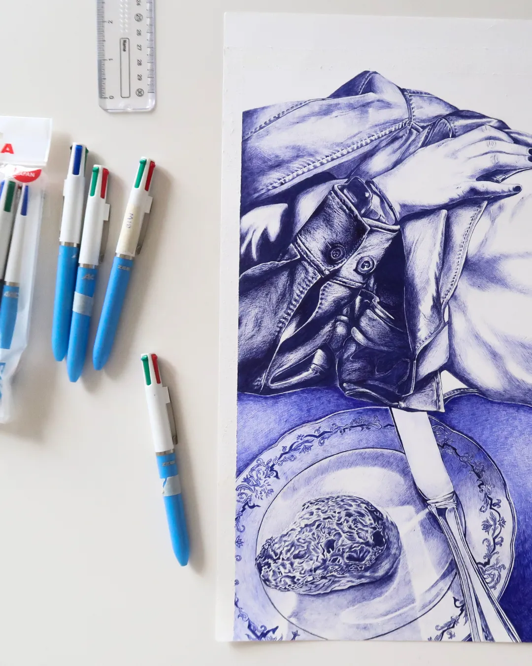 Blue ink drawing of a hand with painted nails resting on a jacket sleeve, above a plate holding a textured object and a knife, with four multicolor retractable blue pens and a clear ruler on a white surface.