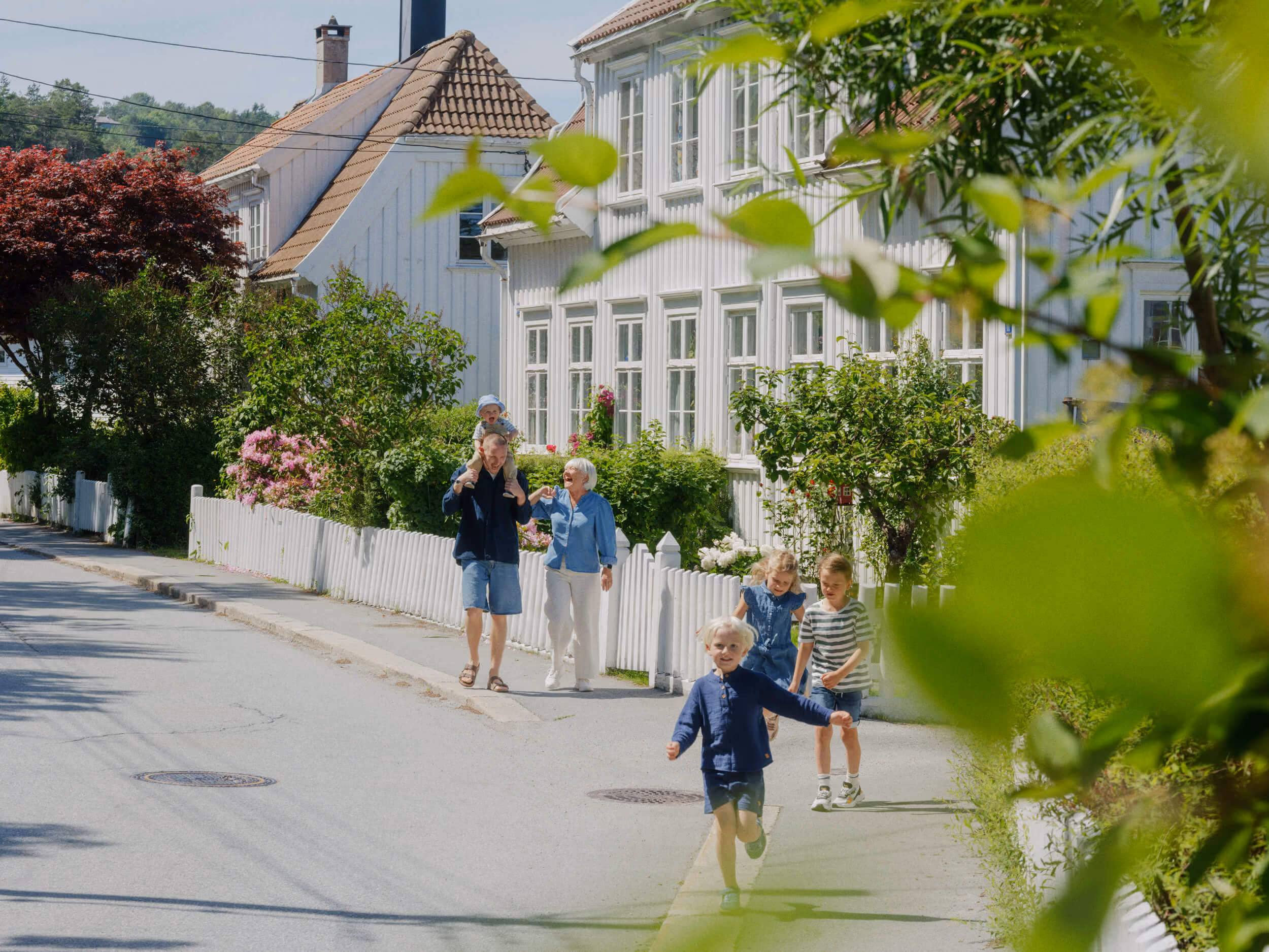 Familie med gamle og små barn som går og løper langs en gate med hvite hus og hage.