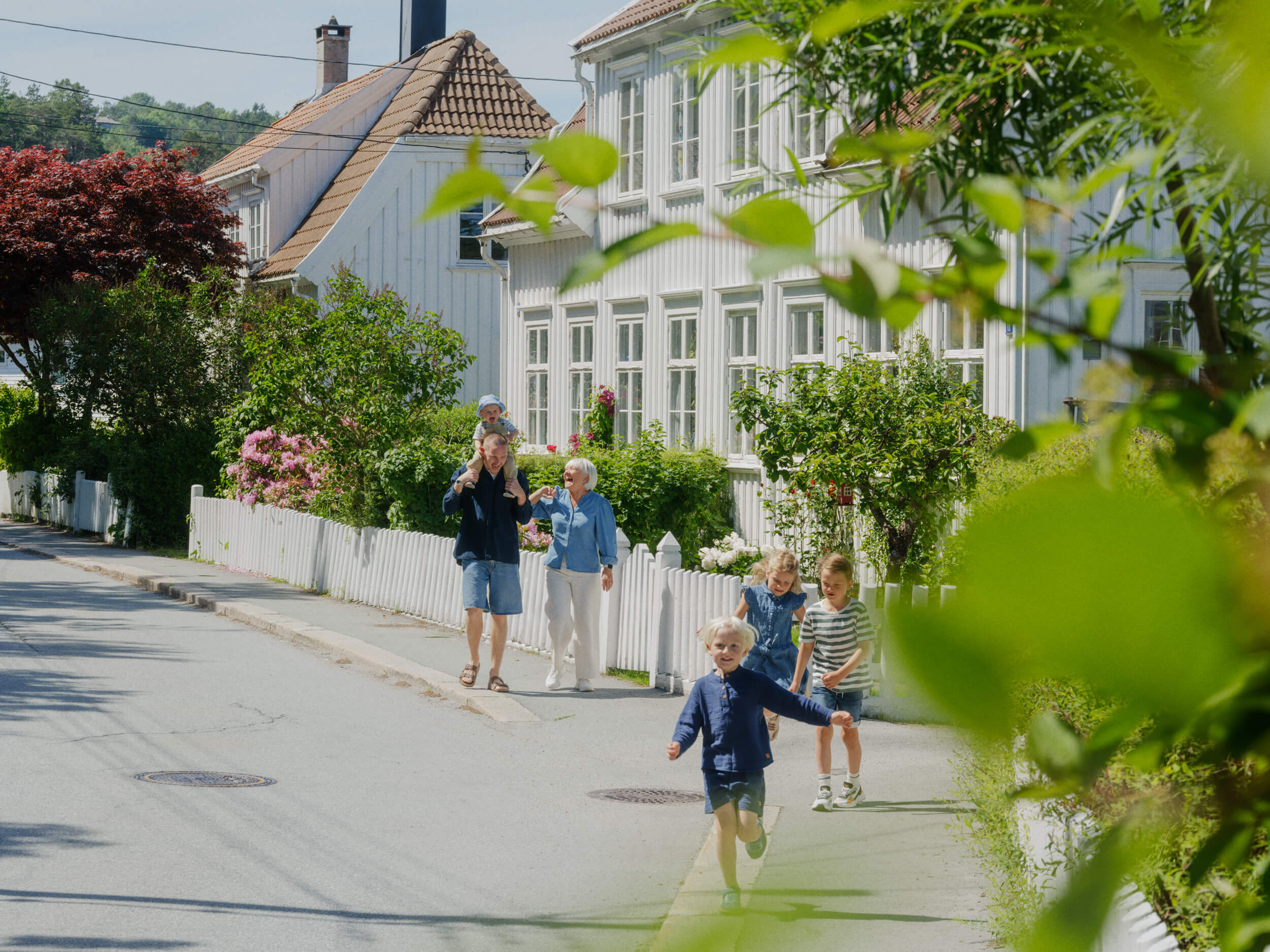 Familie med gamle og små barn som går og løper langs en gate med hvite hus og hage.