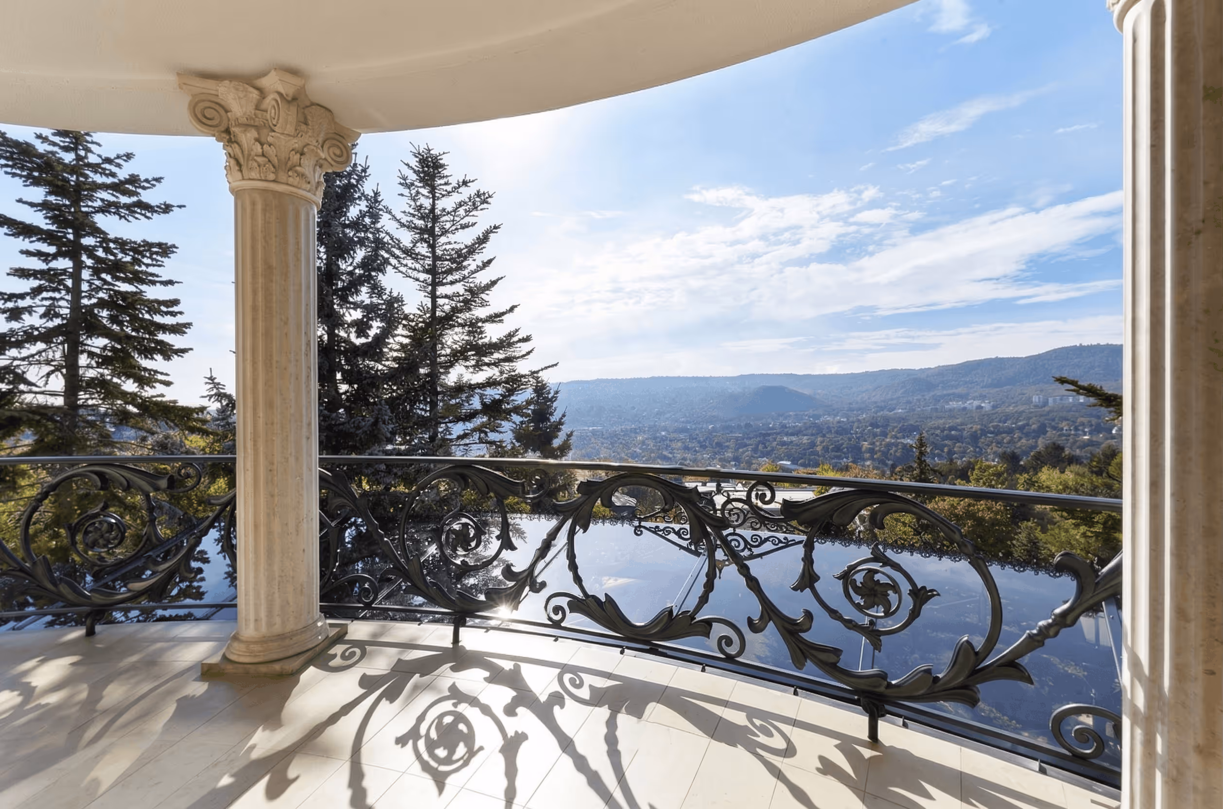 Decorative iron balcony railing casting shadows, with mountain and forest views under a blue sky.